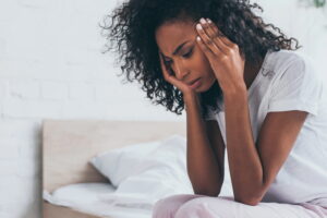 Woman sitting on a bed with hands on her temples, looking stressed or overwhelmed in a bedroom setting.